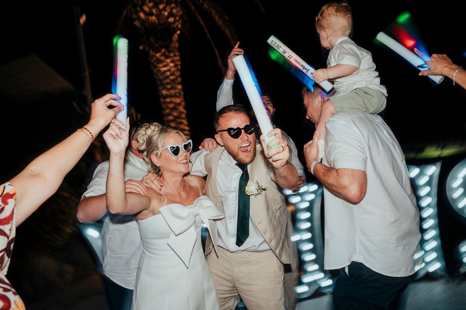 Wedding party celebrates with drinks and laughter at a reception with colorful lighting and floral decor.