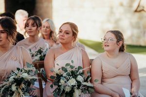 Row of bridesmaids in blush pink dresses sitting together outdoors during a wedding ceremony.