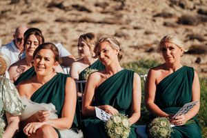 Wedding party in emerald green dresses sits together outdoors on a sunny day, holding white floral bouquets.