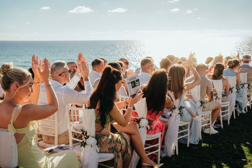 Wedding guests seated at outdoor reception raise their hands in celebration during sunset by the ocean.
