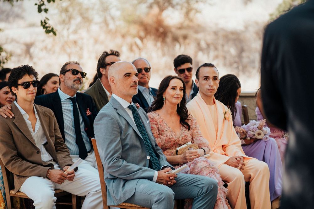 Wedding guests seated outdoors in formal attire watching ceremony during daytime celebration.