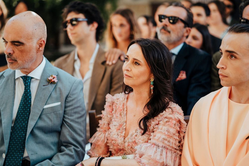 Wedding guests sit together at an outdoor ceremony with soft natural lighting and floral decorations.