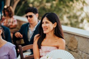 Guests at an outdoor wedding ceremony smile and hold decorative fans on a sunny day.