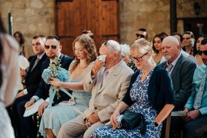 Wedding guests react emotionally during ceremony while sitting in wooden chairs at outdoor venue.