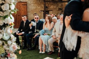 Wedding guests seated outdoors in garden chairs watching emotional ceremony under string lights and floral decor.
