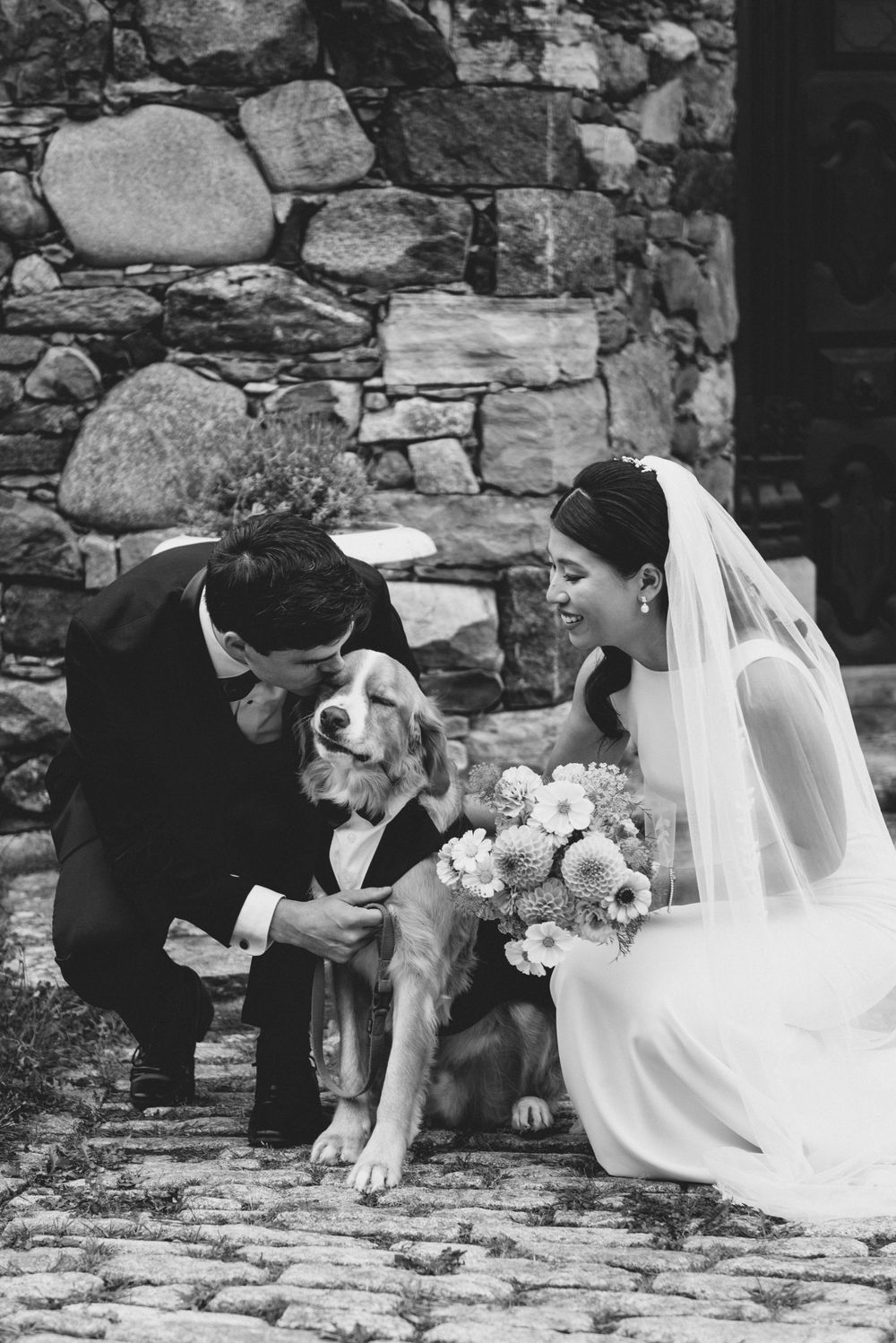 A bride in white dress and groom in suit share a special moment with their dog against a rustic stone wall.