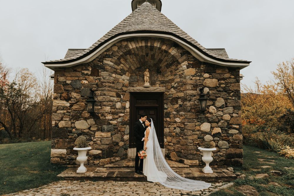 A couple shares a romantic moment outside a rustic stone chapel with autumn trees in the background.