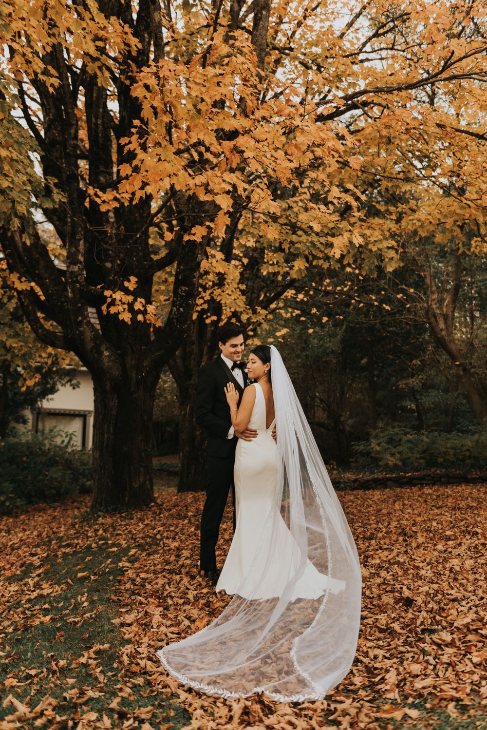 A couple shares a romantic moment under golden autumn trees with fallen leaves and a flowing white wedding veil.