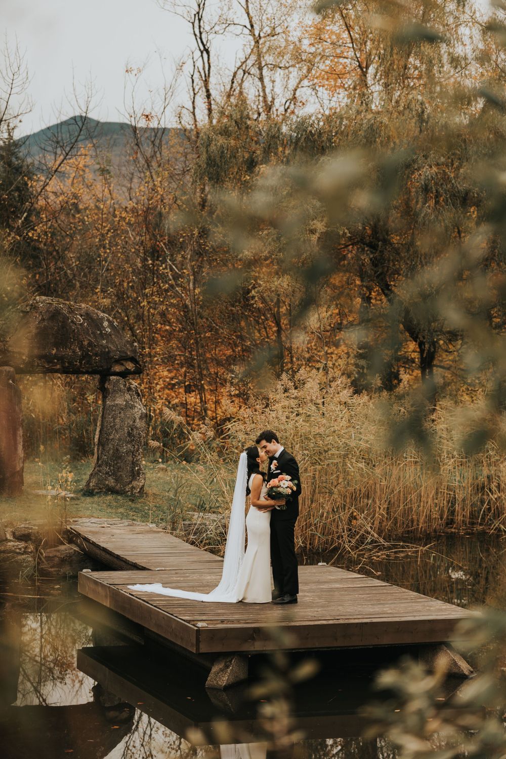 A couple stands together on a wooden dock surrounded by autumn foliage in warm golden light at sunset.