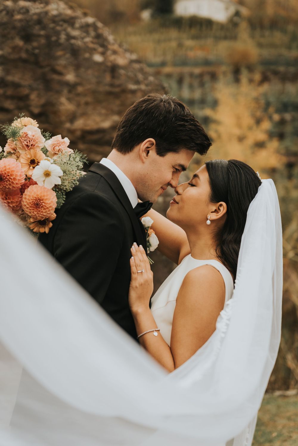 A romantic wedding moment captured with a flowing white veil and peach-colored floral bouquet in a warm autumn setting.