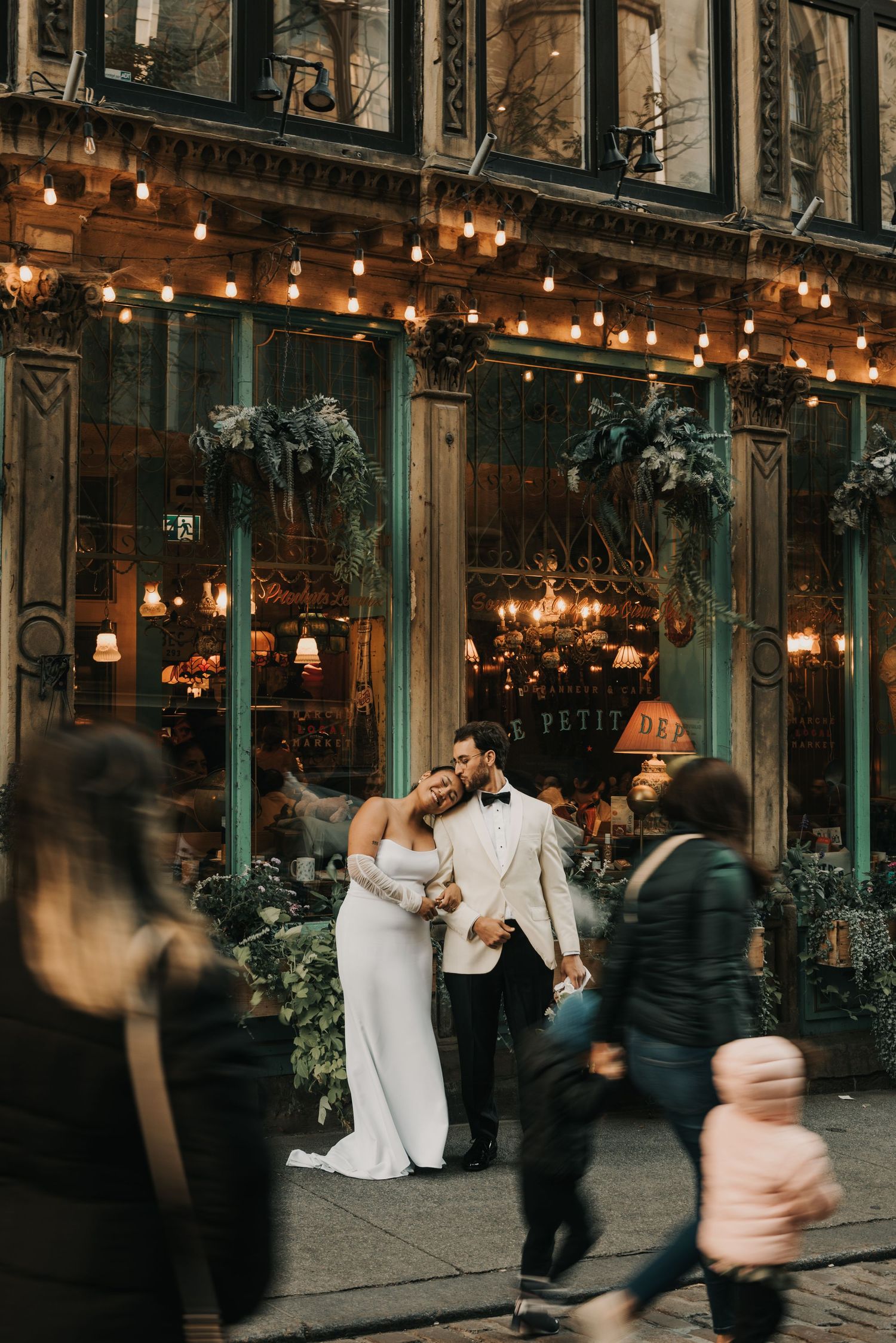 Couple poses outside vintage storefront with glowing string lights and passersby on a cozy evening in the city.