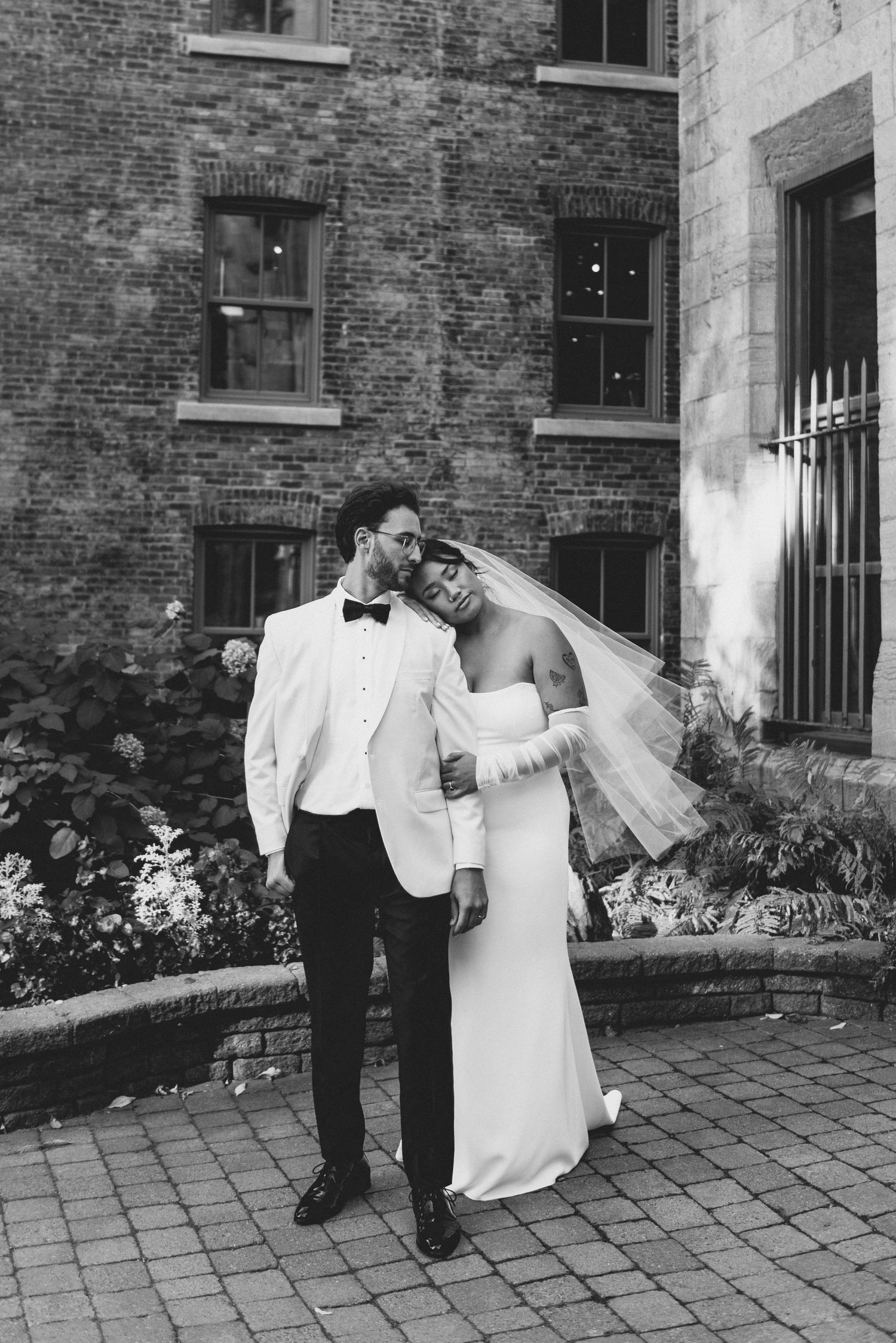 A couple in wedding attire poses against a brick wall background with paved courtyard on a romantic black and white evening.