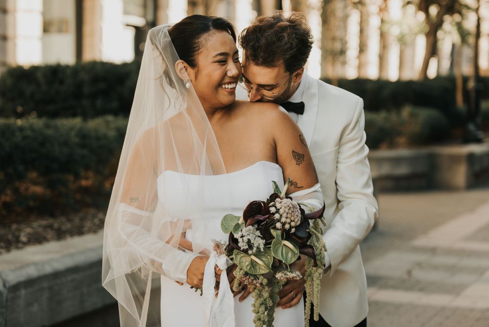 A romantic wedding moment captured in warm sunlight as two embrace, with a beautiful bouquet and flowing veil.