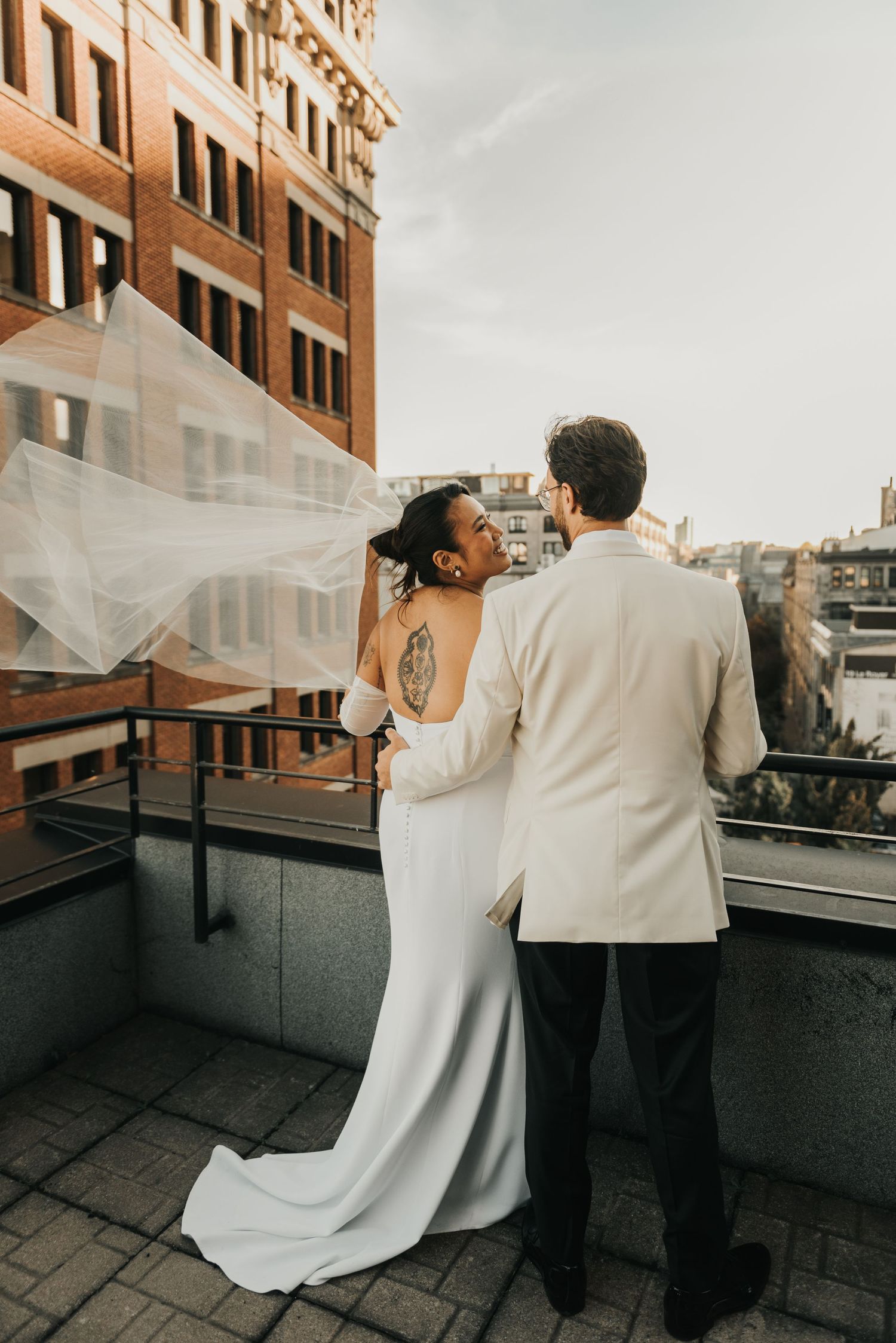 A couple shares an intimate moment on a rooftop terrace, with the bride's veil flowing in the wind against urban architecture.