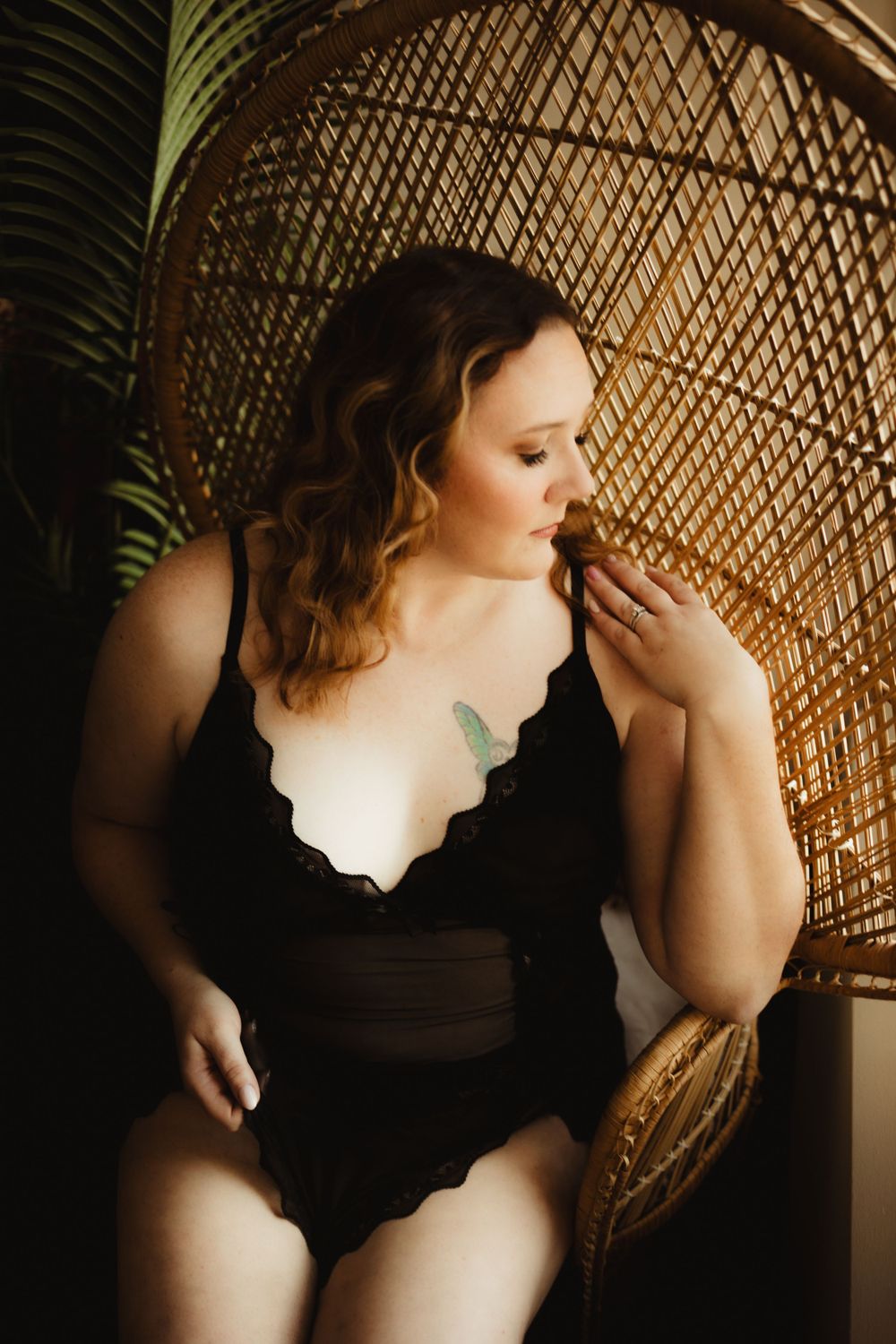 Boudoir photo featuring a black scalloped swimsuit against a woven peacock chair and tropical palm backdrop.