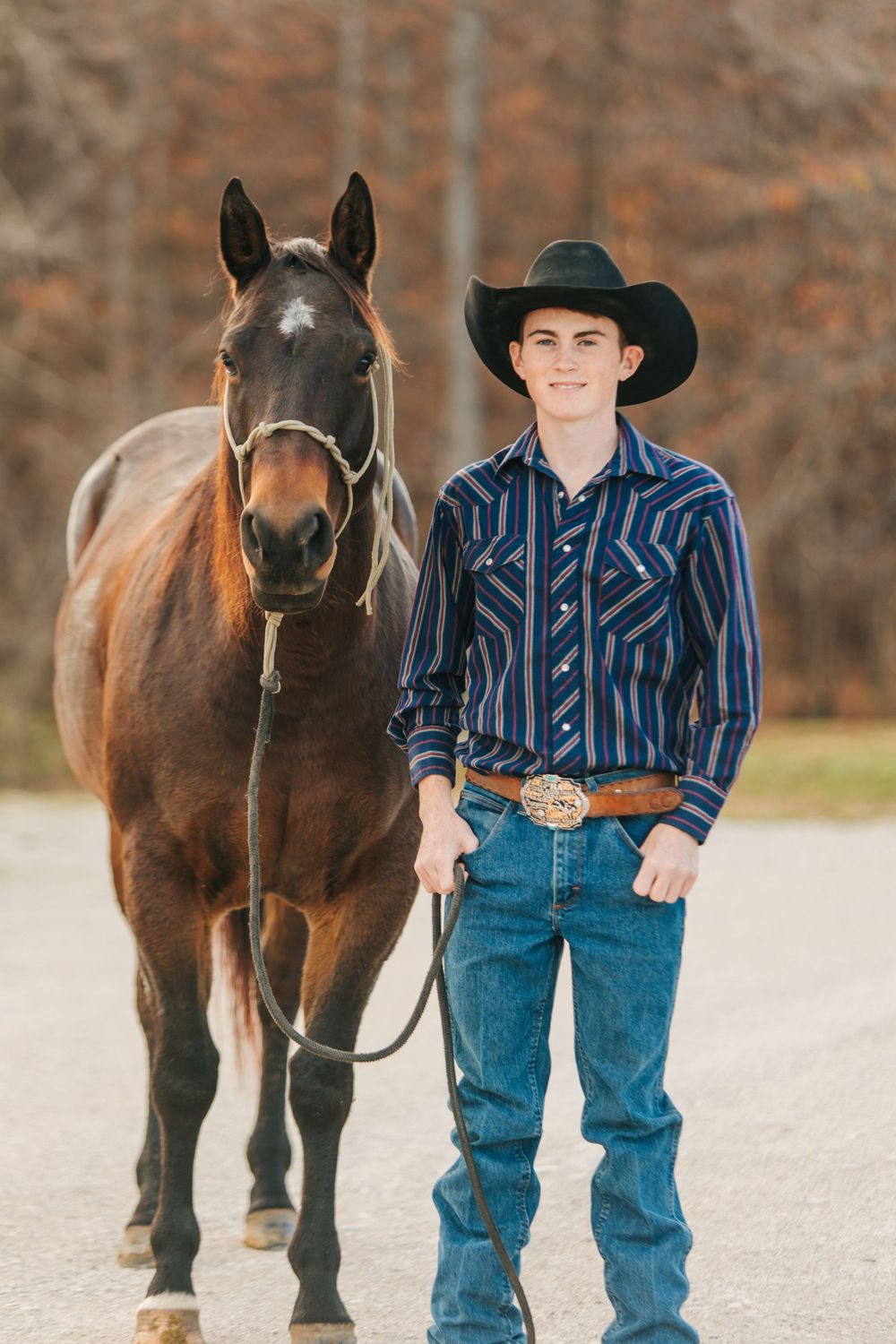 Young cowboy in western wear stands with a dark brown horse on a dirt path in autumn.