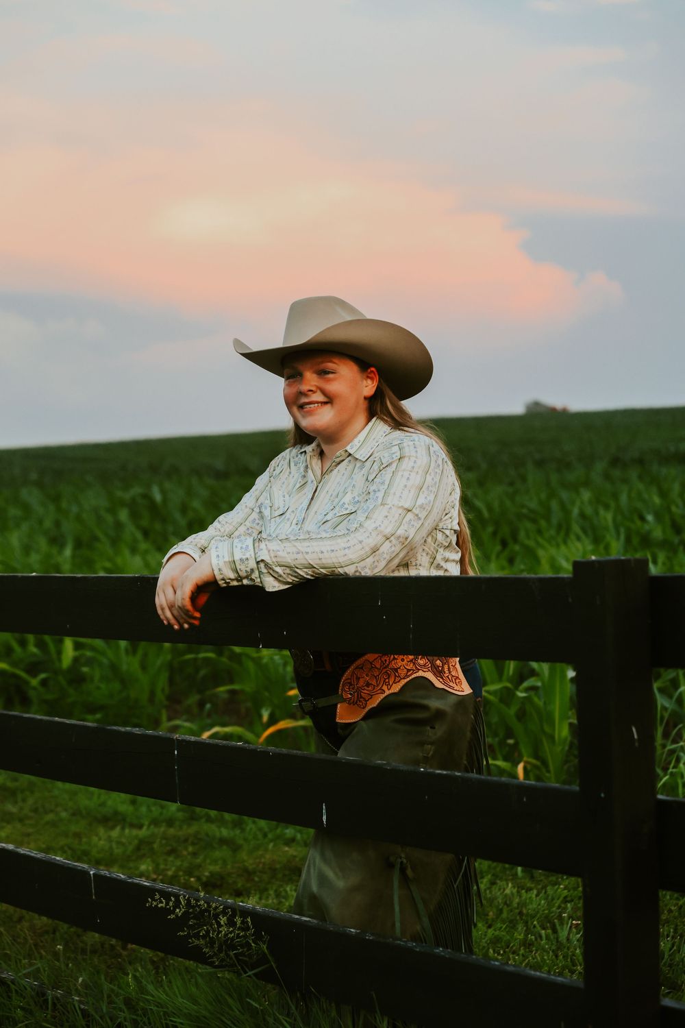 A cowgirl in western attire leans against a black fence at sunset with cornfields in the background.