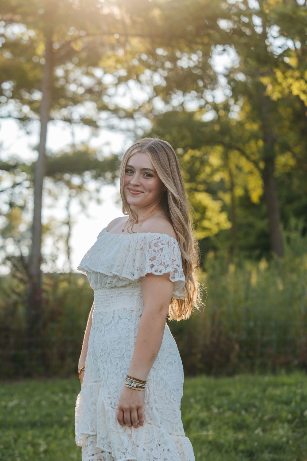 A woman in a white lace dress poses in a sunlit field during golden hour.