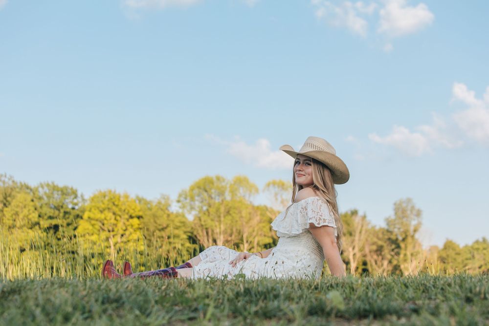 A woman in a white dress and cowboy hat sits peacefully in a grassy field.