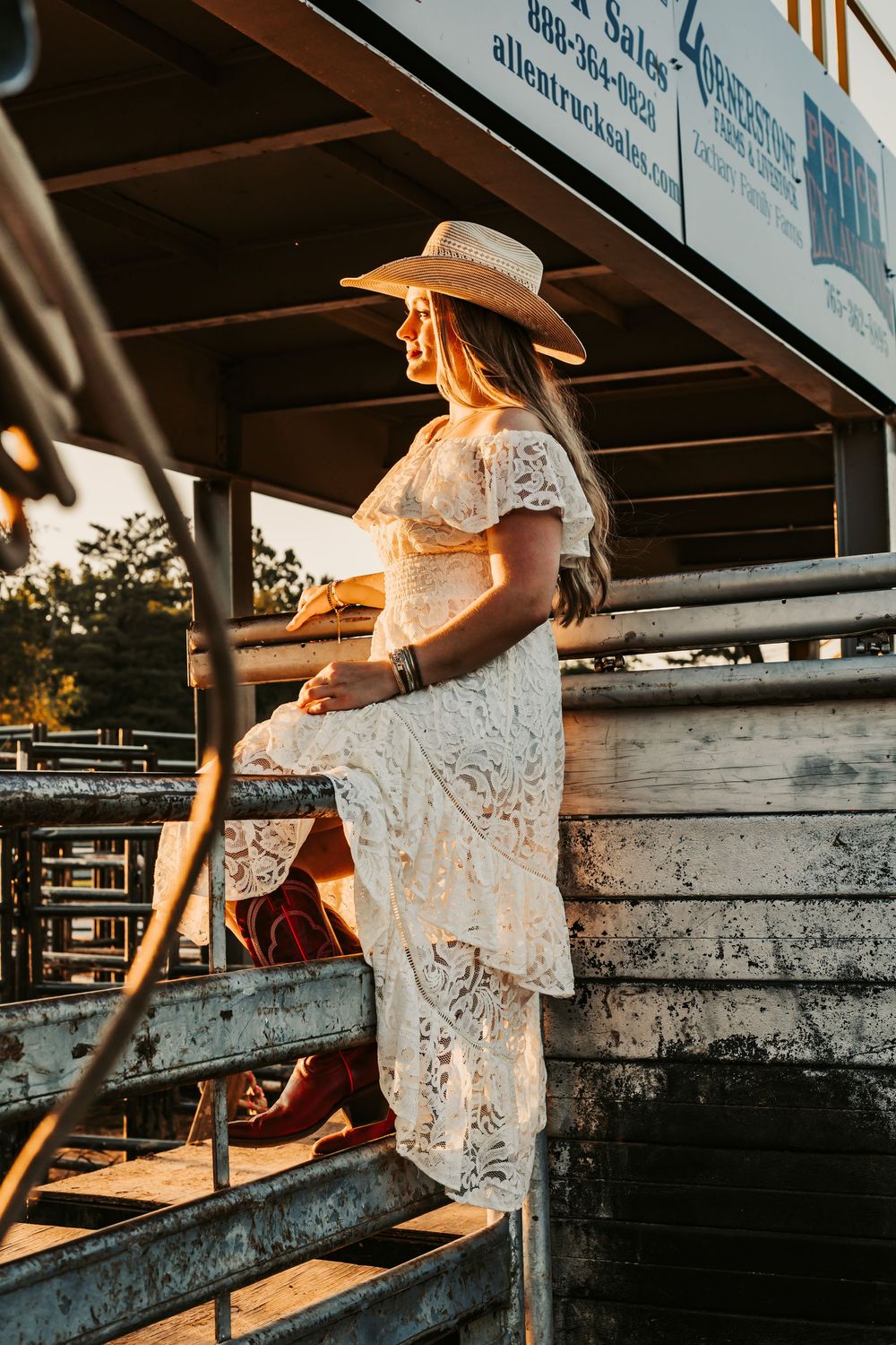 A woman in a white lace dress climbs behind rodeo shoots in warm sunset lighting.