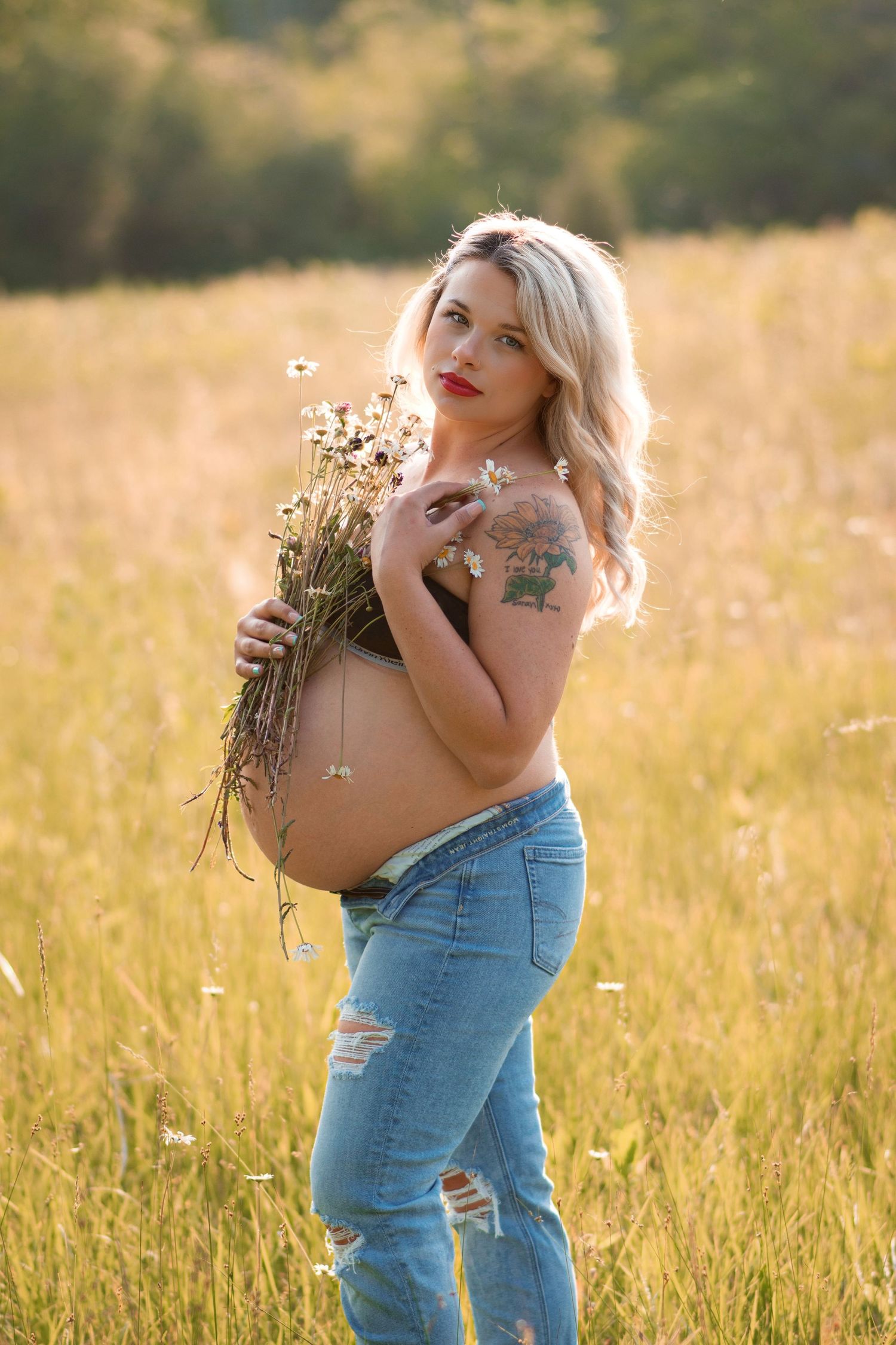Pregnant blonde woman holding wildflowers in a golden sunlit field wearing ripped jeans.