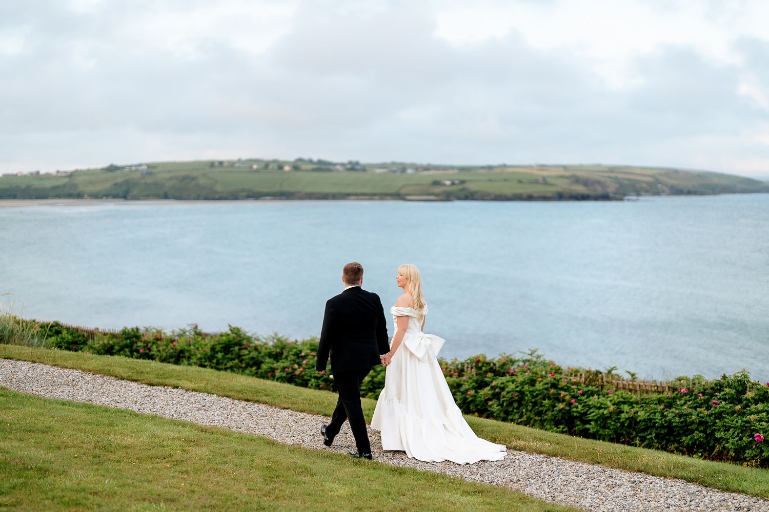 A couple in formal wedding attire walks along a coastal path overlooking a serene bay with green hills in the distance.