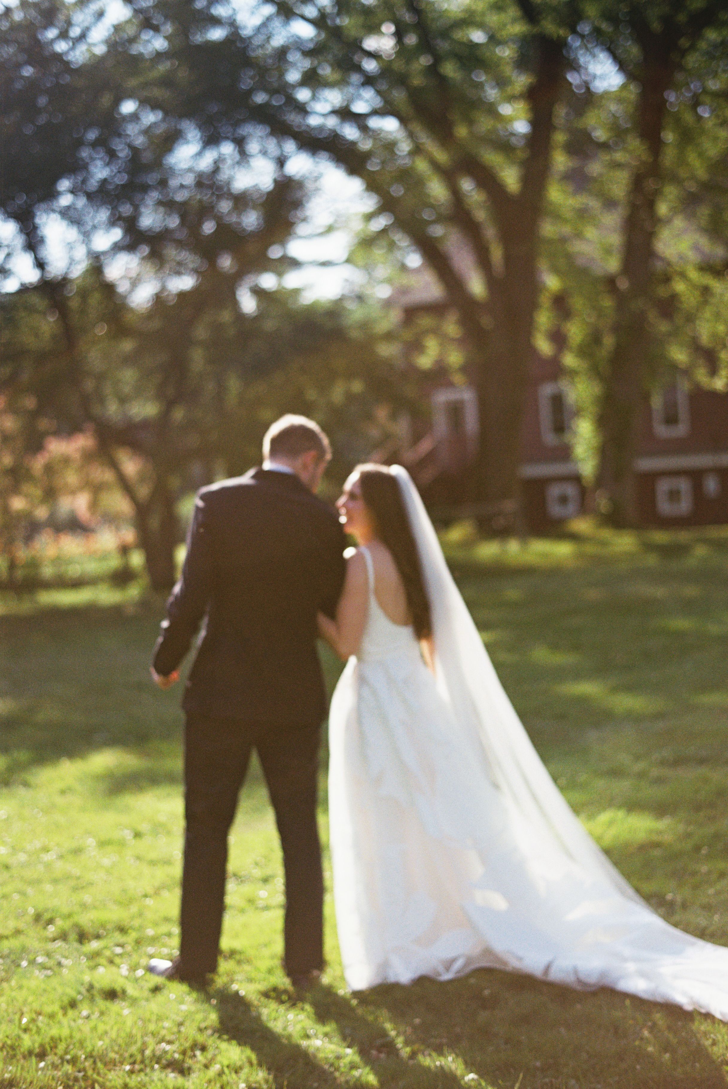 Bride and groom walking hand in hand on lush green lawn with red barn and trees in golden sunlight.