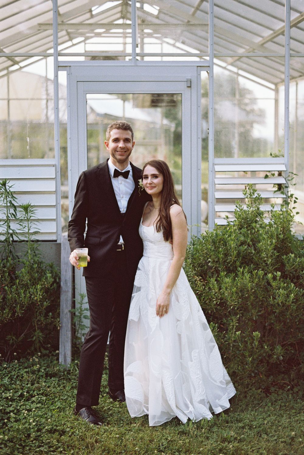 A bride in a white gown and groom in a black tuxedo pose together in front of a glass greenhouse.