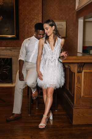 Couple posing in a wood-paneled room, a woman in a white beaded feather-trim dress and heels leaning against a seated man in cream attire.
