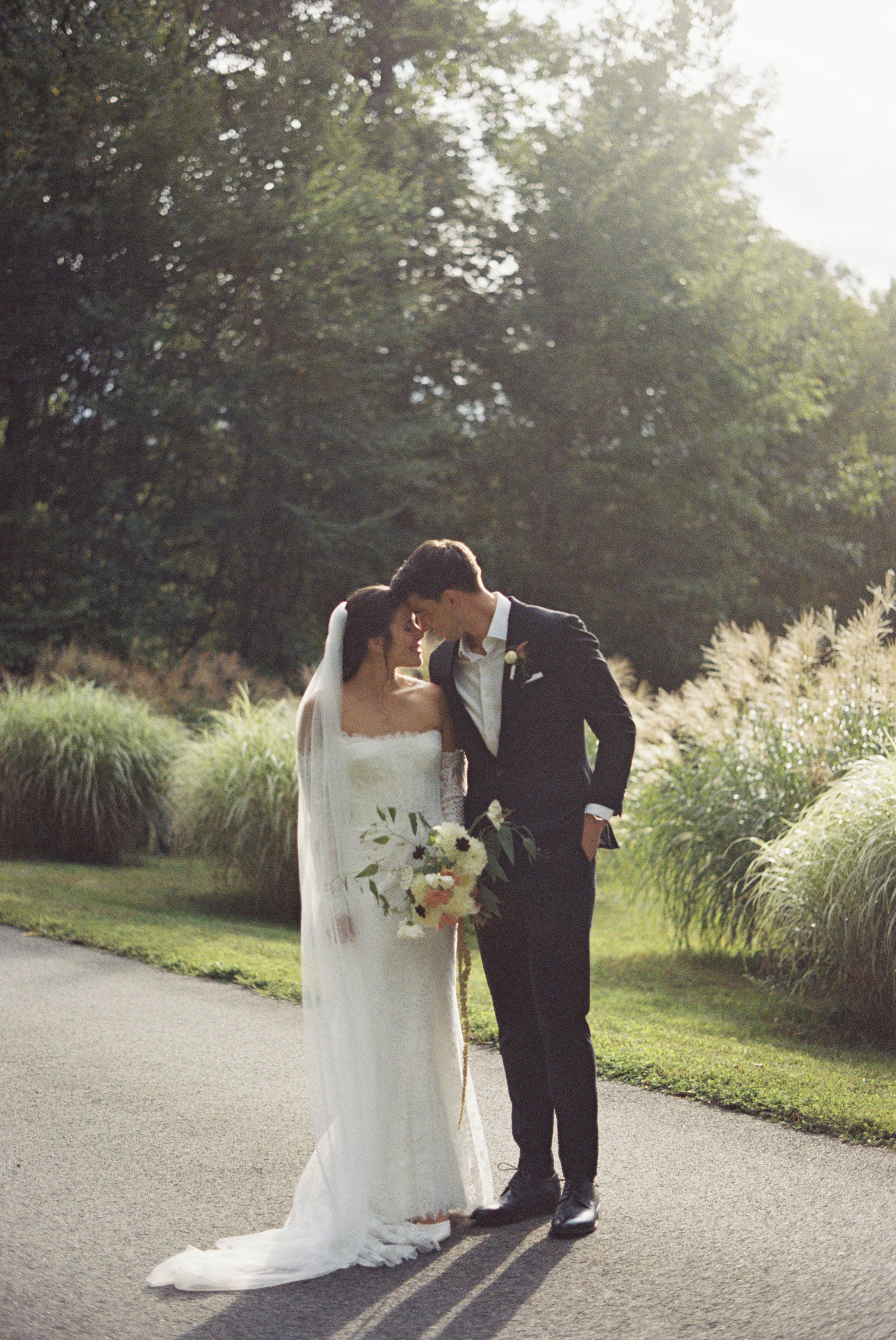 A 35mm film portrait of bride and groom sharing an intimate moment outdoors on a garden path surrounded by lush greenery and trees.