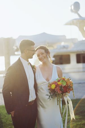 Bride and groom smile together outdoors near a yacht, she holds a vibrant red and yellow bouquet on their wedding day.