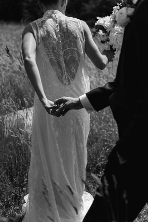 Black and white photo of bride in lace wedding dress holding hands with groom in a field.