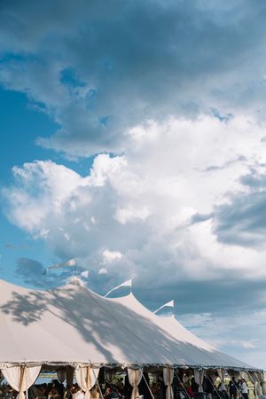 White event tent peaks against a dramatic blue sky with billowing clouds at an outdoor gathering.