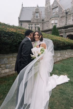 Smiling bride in a white gown and long veil holding a bouquet as her groom in a black suit kisses her cheek outside a stone mansion.
