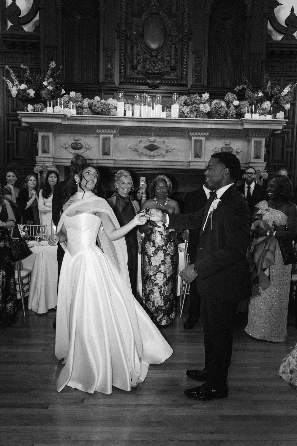 Black and white photo of a bride in a satin gown and groom in a suit sharing a first dance as wedding guests watch in an ornate hall.
