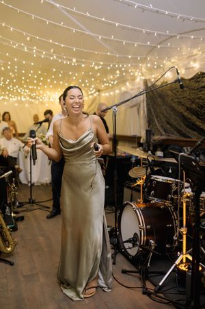 Smiling female vocalist in a satin sage green gown sings at a wedding reception under a tent with string lights and a live band.