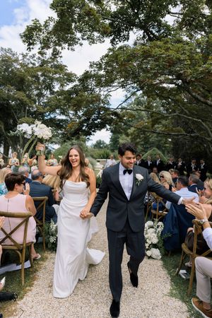 Joyful bride and groom walk down the aisle after wedding ceremony outdoors, surrounded by cheering guests.
