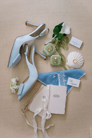 Light blue bridal heels with bow, green velvet ring box, diamond ring, seashell, floral accents, and elegant stationery on linen backdrop.