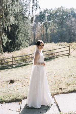Bride in white gown and veil smiling outdoors near wooden fence and trees on sunny autumn day.
