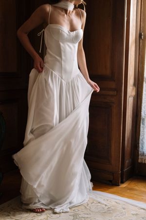 A white wedding dress with a corset bodice and flowing skirt stands elegantly near wooden doors in natural light.