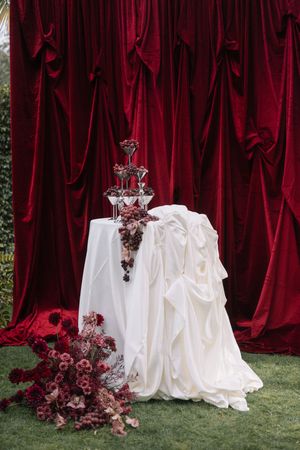 Elegant wedding decor with draped red velvet curtains, white tablecloth, and cascading burgundy floral arrangements.