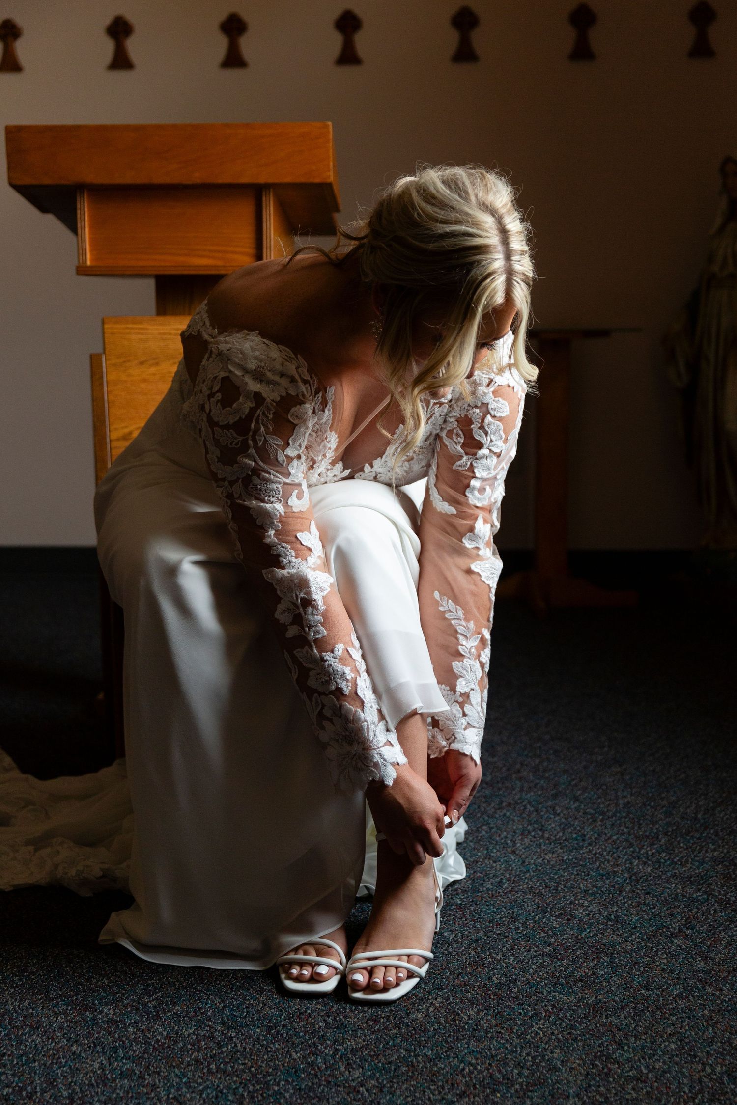 Bride in long sleeve lace gown adjusting white sandals while seated on wooden church pew.