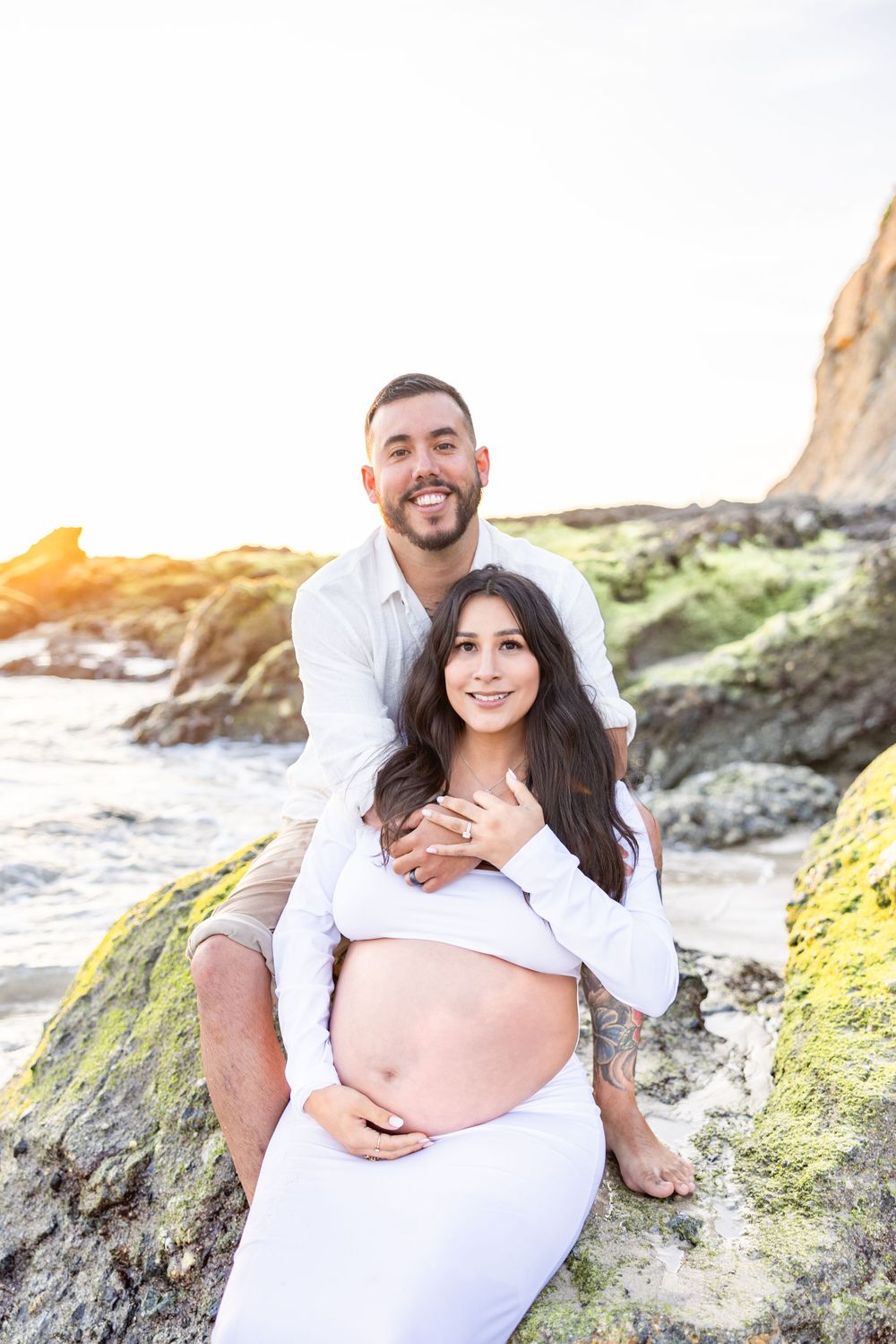 Expectant couple posing together on rocky beach during golden hour maternity photoshoot.