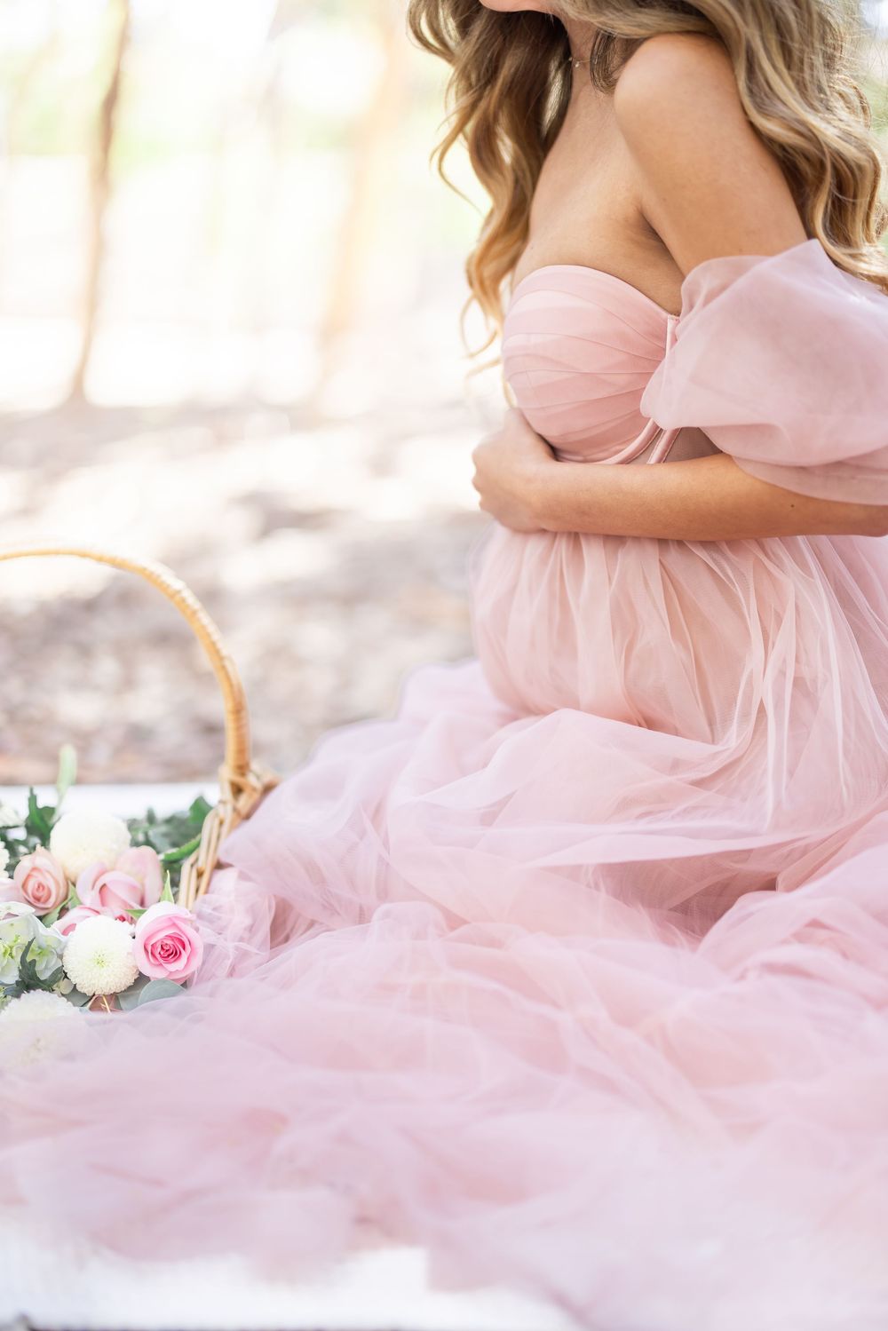 Pregnant woman in elegant pink tulle off-shoulder gown sitting outdoors near a wicker basket with pink and white roses.