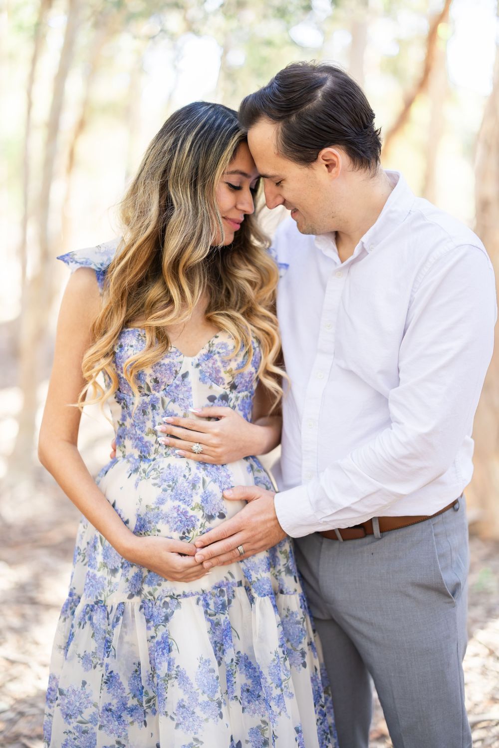 Couple sharing a tender moment outdoors, woman in floral maternity dress cradling baby bump, man in white shirt embracing her.
