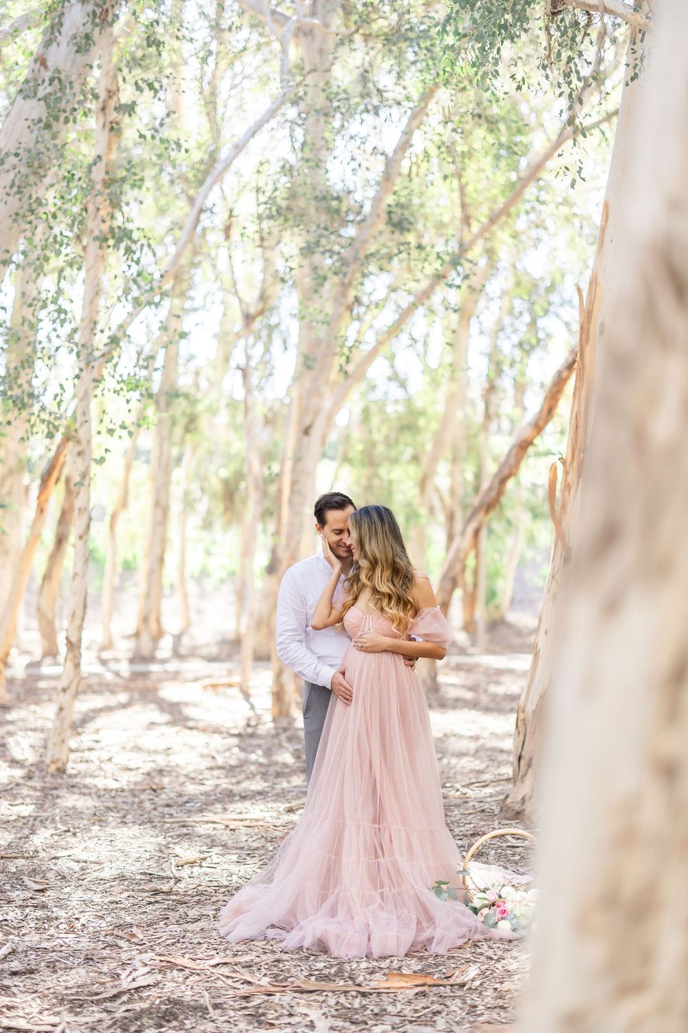Couple embracing in a sunlit forest, woman in a flowing blush pink tulle gown, surrounded by tall eucalyptus trees.