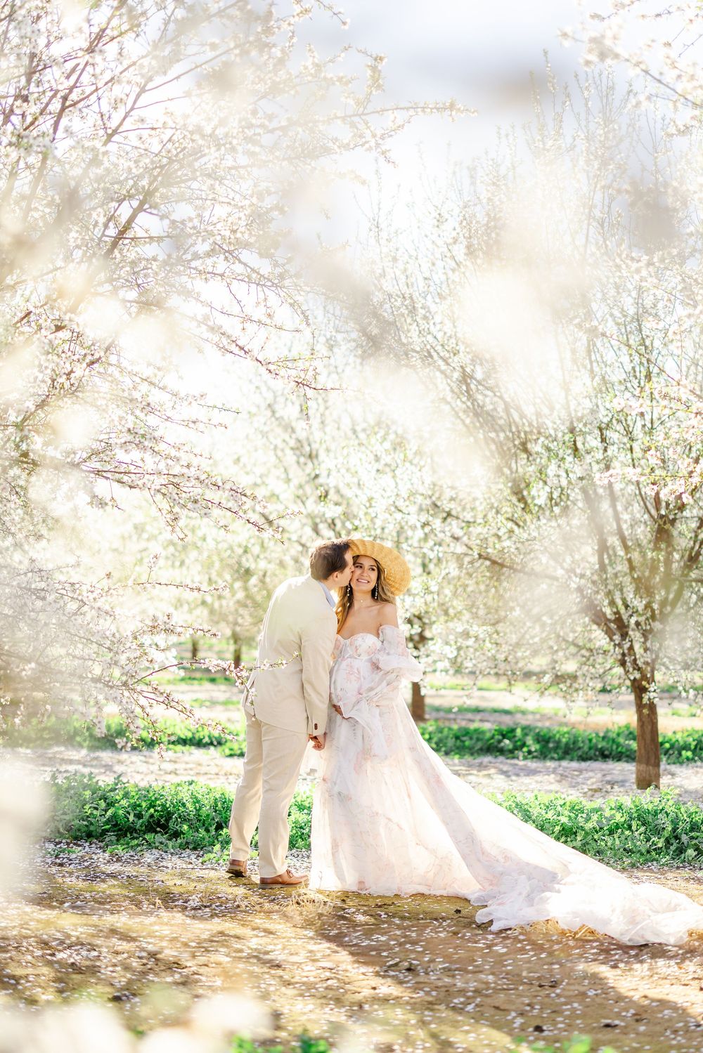 Couple in wedding attire embracing among blooming white trees in a sunlit outdoor garden setting.