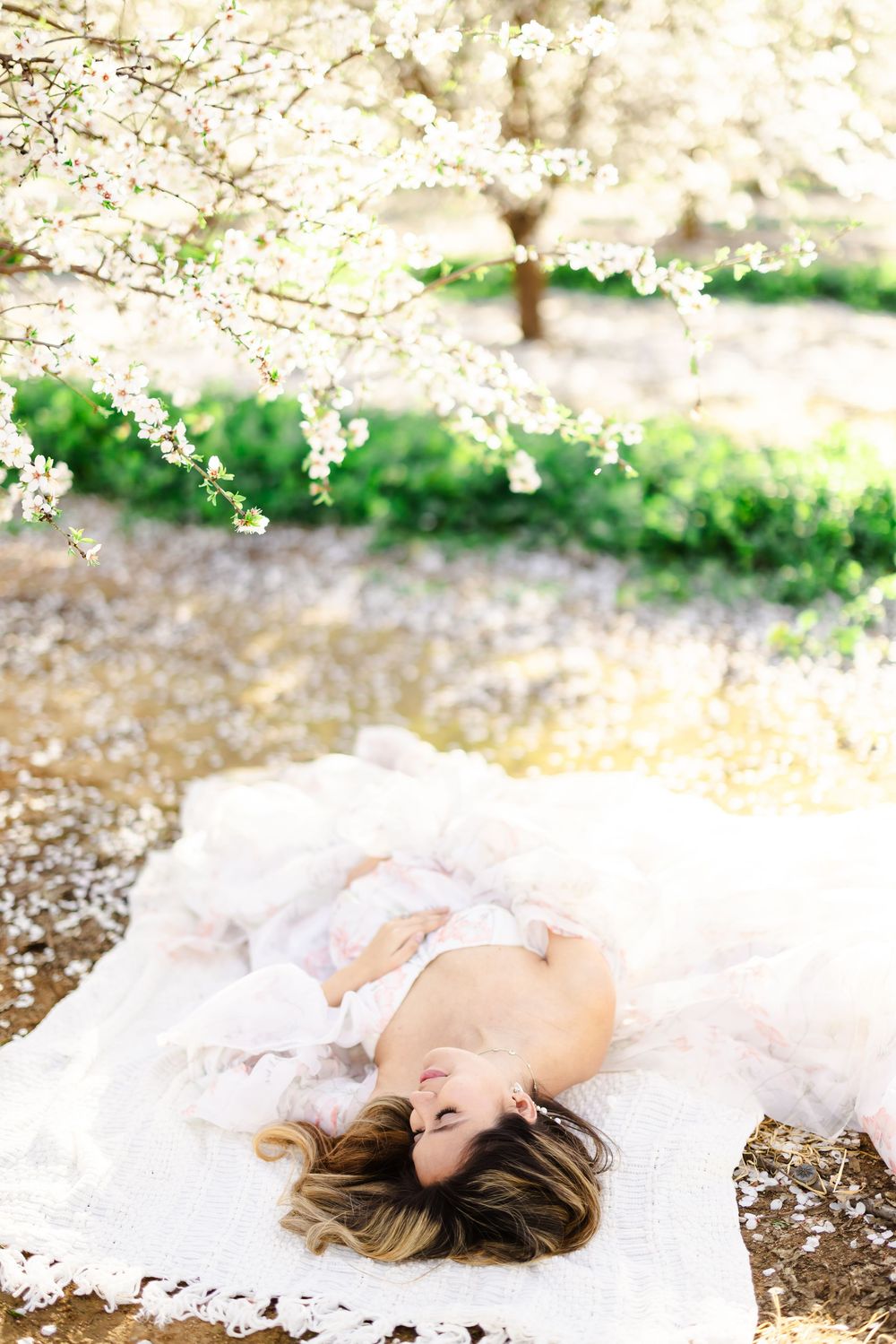 Woman in white dress lying peacefully on a blanket under blooming cherry blossom trees in spring.
