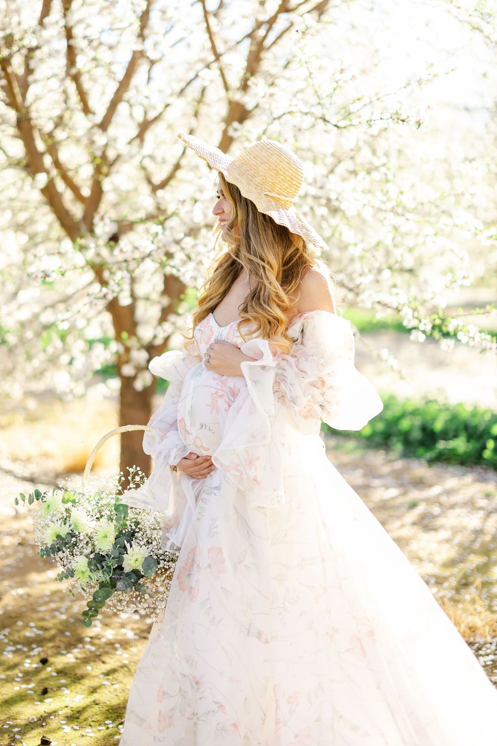 Woman in floral off-shoulder gown and straw hat holds bouquet among blooming white trees in soft sunlight.