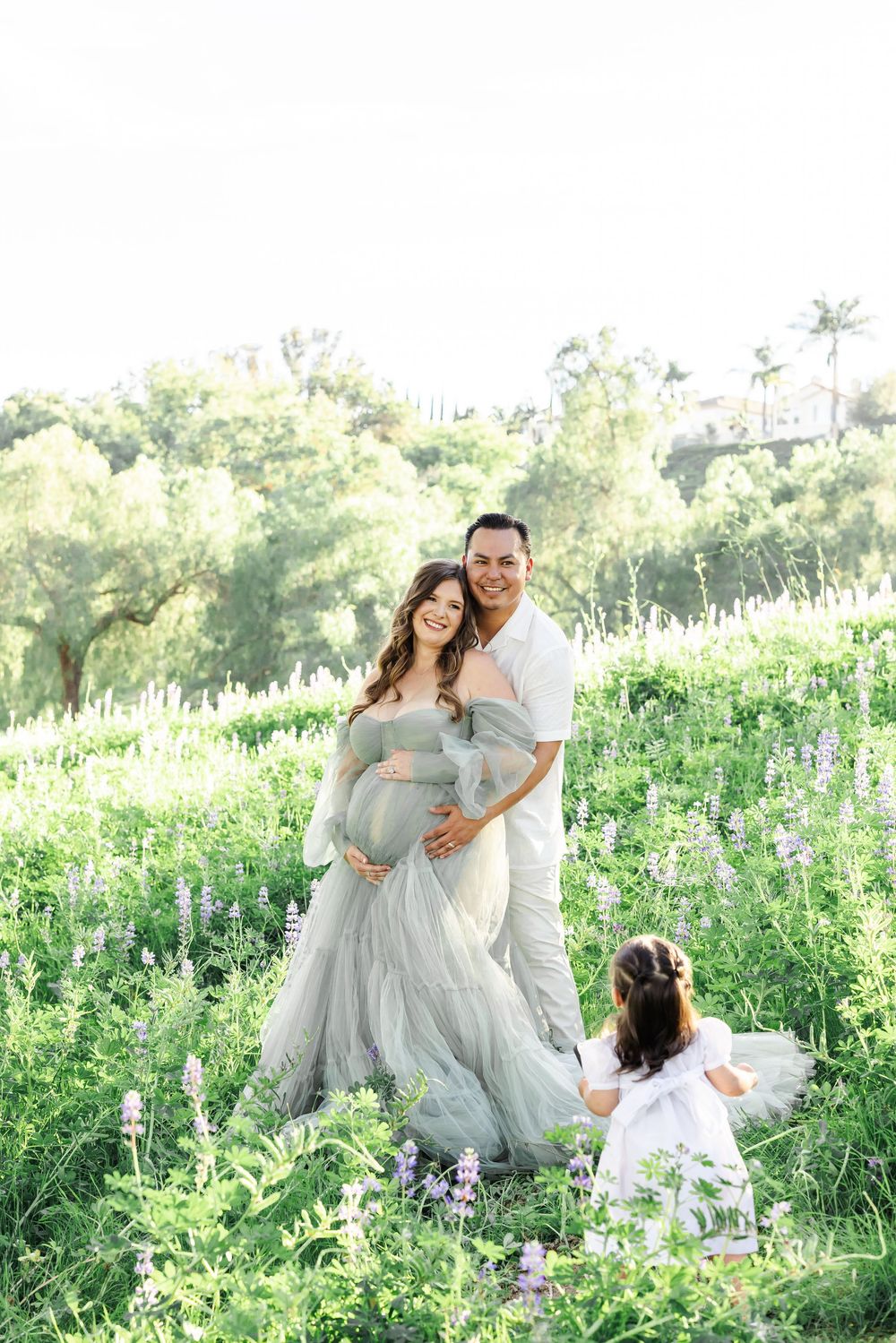 Pregnant woman in sage tulle gown with husband and daughter in a wildflower field during a maternity photoshoot.